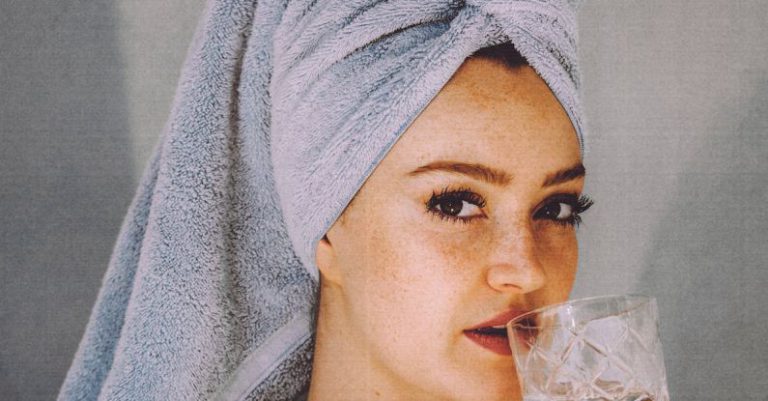 Clean Beauty - Young woman with hair wrapped with towel and bared shoulders drinking water and looking at camera